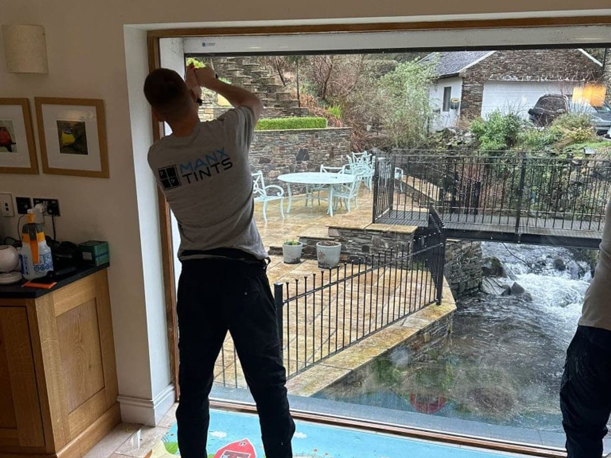 A ManxTints installer fitting film to a large window above a stream